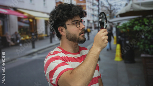 Billede på lærred Young man with beard examines street scene using magnifying glass, wearing striped shirt outdoors, showcasing curiosity and exploration in urban environment