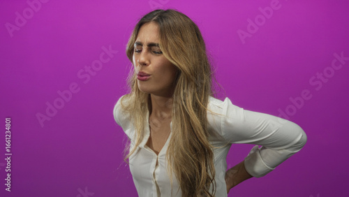 Young blonde woman wearing white blouse holds lower back in purple studio setting; pain discomfort backache fatigue.