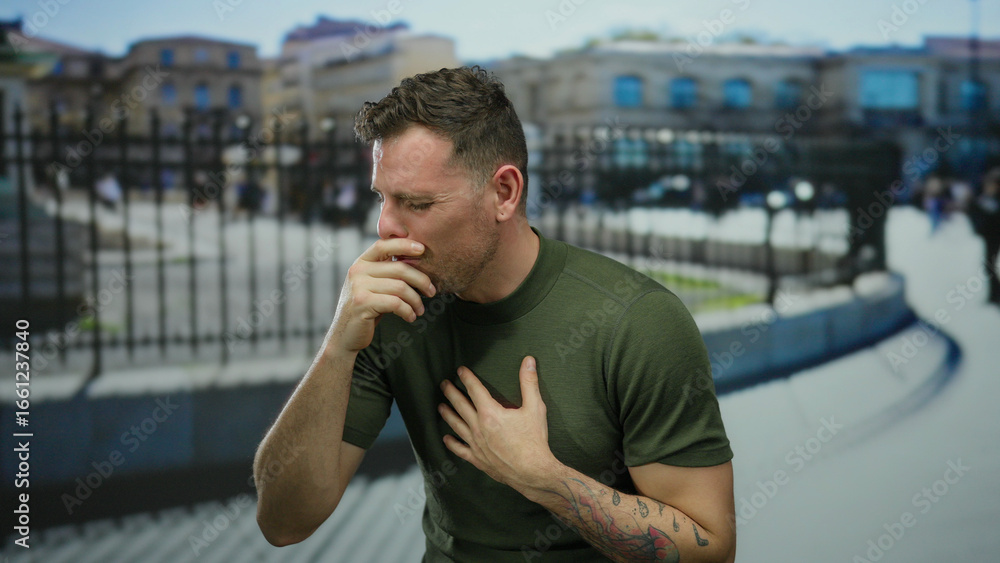 Fototapeta premium Young man sneezing outdoors in urban area wearing green shirt with tattoos visible against blurry city background during day.