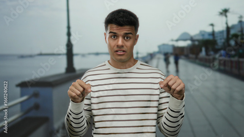 Young man gestures with expression on a seaside promenade showing a relaxed and humorous demeanor captured against a serene beach background.