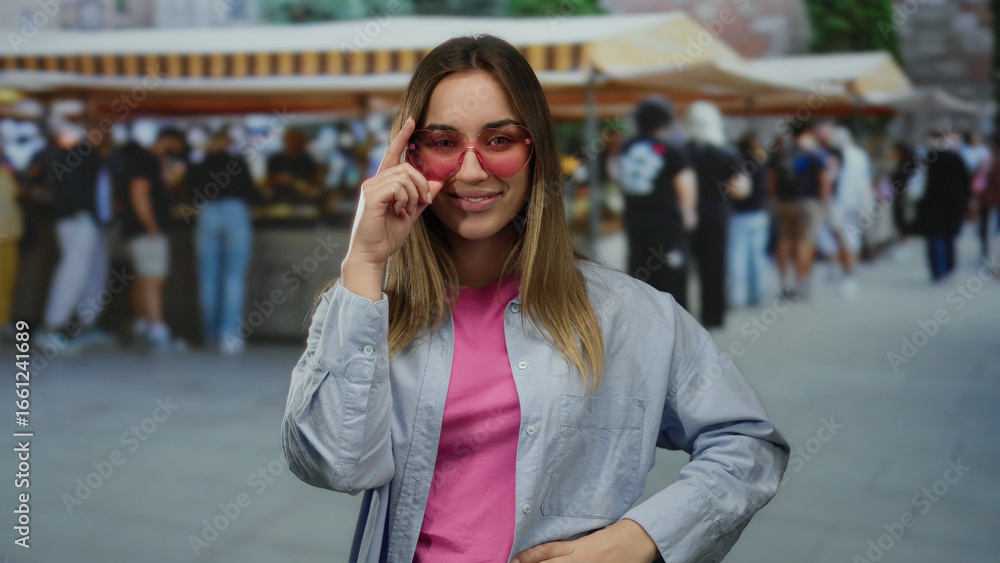 Fototapeta premium Woman in pink sunglasses poses confidently at an outdoor market, capturing a lively street scene with people in the background, emphasizing a vibrant shopping atmosphere.