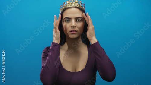Photos Young hispanic woman wearing a crown standing against a vibrant blue wall with a confident expression, showcasing elegance and strength