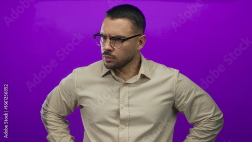 Photos Young hispanic man wearing glasses pointing finger forward in vibrant purple studio; determination focus authority