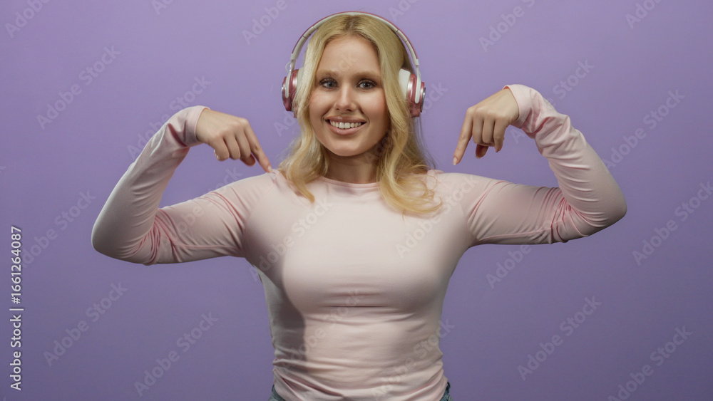 Fototapeta premium Young blonde woman wearing headphones against a purple background smiling and pointing downwards with both hands in a playful manner