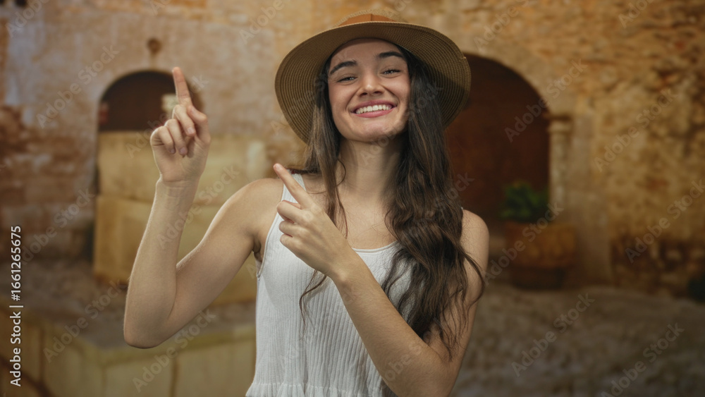 Naklejka premium Teenage woman wearing straw hat and white tank top points finger to sunlit street in old town; adventure joy.