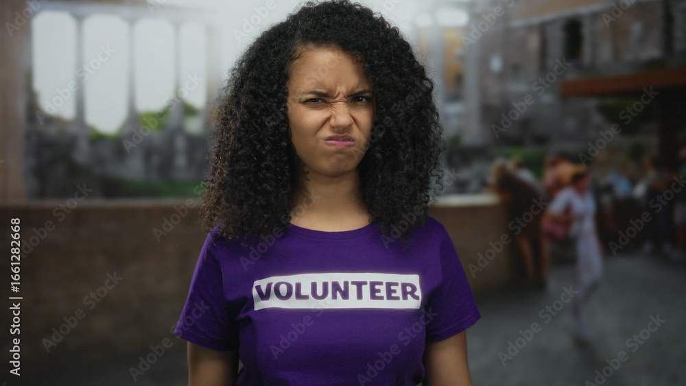 Fototapeta premium Hispanic woman in volunteer shirt stands outdoors on historic town street with thoughtful expression, blending modern engagement with ancient architecture.