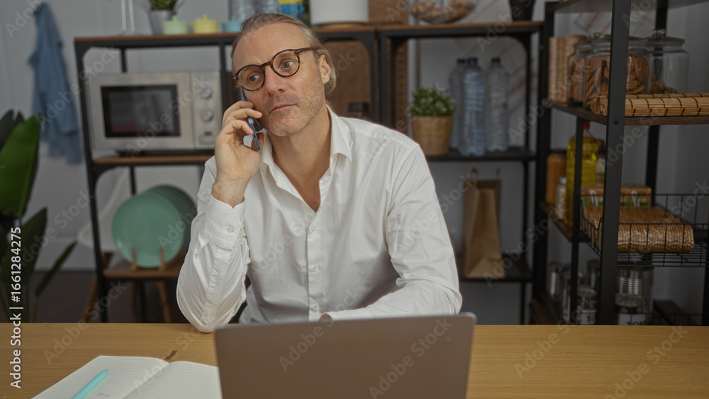 © Krakenimages.com - Mature blonde man talking on cellphone in office with laptop open, conveying business environment with shelves and kitchenware in background, portraying focus and engagement. © Krakenimages.com - Mature blonde man talking on cellphone in office with laptop open, conveying business environment with shelves and kitchenware in background, portraying focus and engagement.