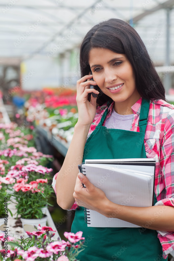 Fototapeta premium Female horticultural worker talking on smartphone while holding notebooks in greenhouse, copy space
