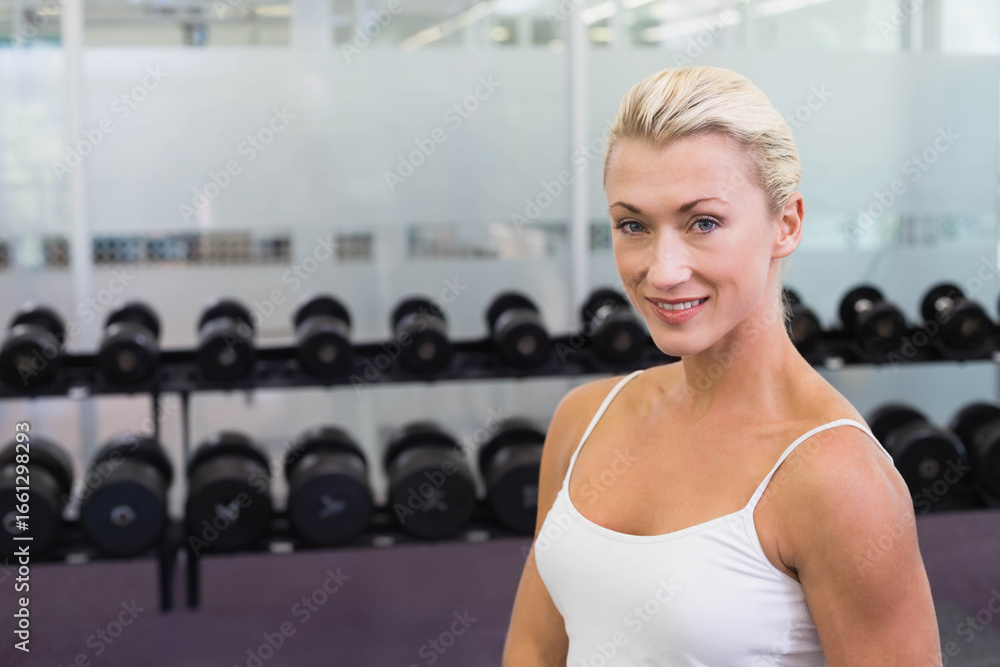 Fototapeta premium Woman standing in gym wearing white tank top and leggings by dumbbell rack, copy space