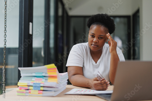African businesswoman experiencing headache and stress while working on a laptop and documents in a modern office, dealing with chronic pain and seeking medical help