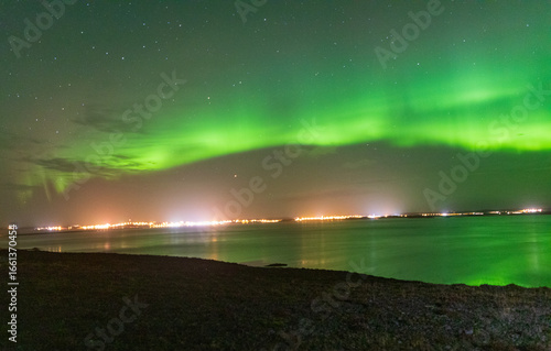 Wallpaper Mural View of shimmering green aurora borealis dances above the distant city lights, mirroring in the tranquil, dark waters, a celestial ballet of light and nature, Vik, Iceland. Torontodigital.ca