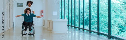Two young friends enjoying playful wheelchair ride in modern hallway with large windows and natural light