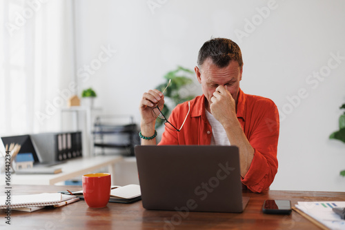 Stressed mature entrepreneur taking off glasses and rubbing his eyes while working on laptop at home, suffering from dry eye syndrome or eye strain after long computer work