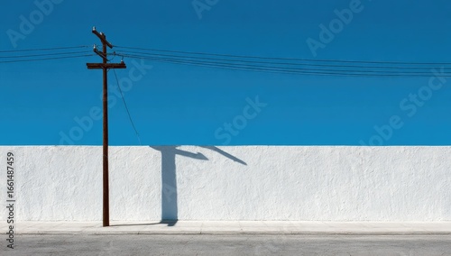 Simple power pole against a vibrant blue sky.  A stark white wall extends below and to the right.  A shadow is cast onto the wall