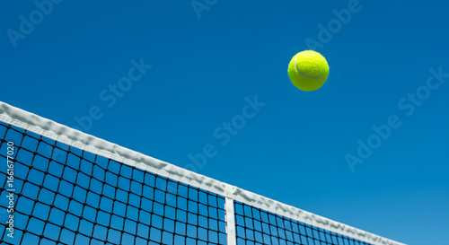 Tennis Ball in Flight: A vibrant tennis ball soars gracefully against a backdrop of a clear, cerulean sky, captured mid-air. The net subtly frames the shot.