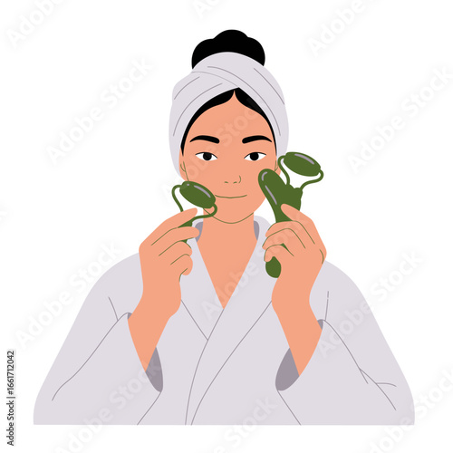 A young woman in a white bathrobe and head towel uses two green jade rollers on her face, embracing a relaxing and mindful beauty ritual.