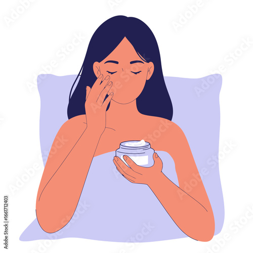 A calm young woman, resting in bed, thoughtfully applies a soothing face cream from a jar, embracing her evening beauty and self-care ritual.