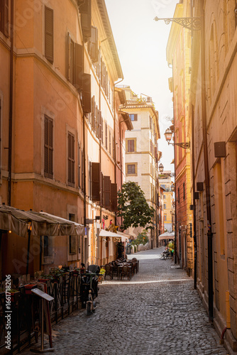 Narrow cobblestone street in Trastevere, Rome, Italy, with cafes, tables, warm sunset light and colorful buildings