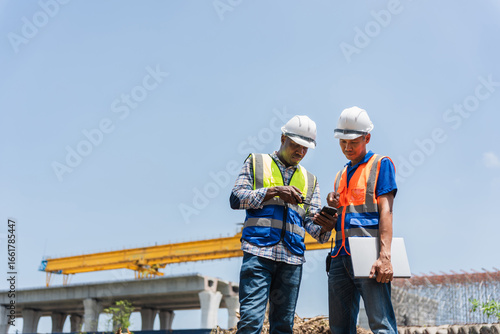Engineers in hard hats and vests looking at a smartphone, Construction workers reviewing project plans on construction site, Civil engineers working at a new road construction site