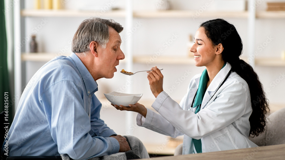 Fototapeta premium Caregiver feeding elderly man in wheelchair at home. Older male patient with restrained movement after stroke eating with caregiver's help. Medical assistance for senior people with partial paralysis