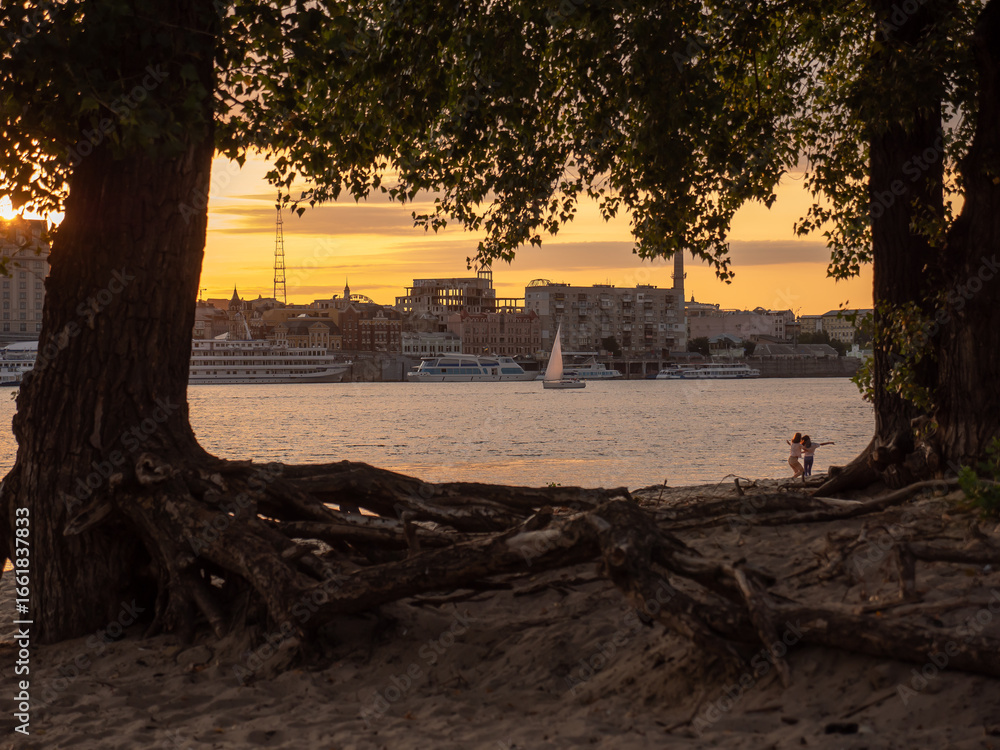 Obraz premium Children playing at an urban sand beach with old trees at sunset on the right bank of the Dnipro river in Podil district in Kyiv, Ukraine. Ancient hill with church, old building and boats in the port