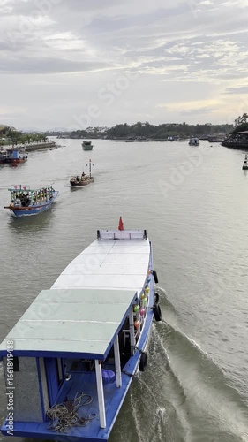 From an elevated perspective, various boats, including tour vessels and local transport, navigate a wide river in Vietnam under a serene, cloudy summer sky. 