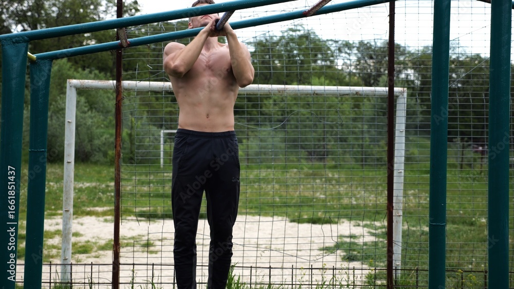 Fototapeta premium Young muscular sportsman doing pull ups at sports ground. Athletic guy exercising outdoor. Strong sporty man working out at summer nature. Concept of healthy and active lifestyle. Slow motion