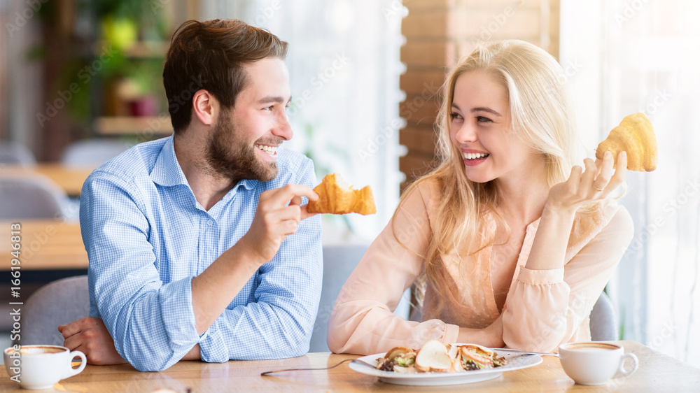 Obraz premium Positive young couple eating healthy breakfast together at coffee shop. Handsome millennial man having dinner with his girlfriend at cafe, enjoying yummy meal, spending happy time with beloved woman