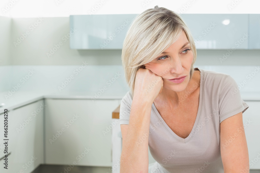 Mature woman sitting at glossy countertop leaning chin on hand gazing left in kitchen, copy space Mature woman sitting at glossy countertop leaning chin on hand gazing left in kitchen, copy space