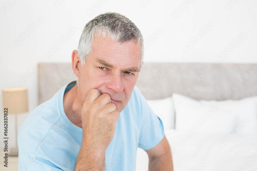 Obraz premium Senior man sitting on edge of bed wearing light blue T-shirt at bedside with nightstand lamp