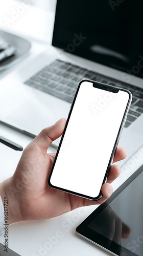 Close-up of a hand holding a smartphone with a blank white screen next to a laptop mobile phone person