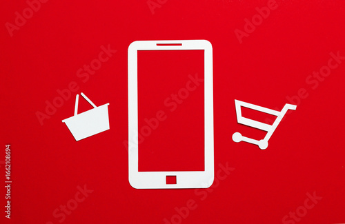 Paper-cut smartphone icon with supermarket basket and trolley on red background. Online shopping