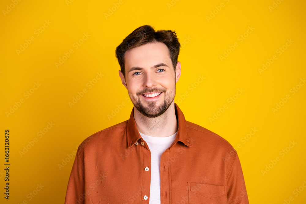 Fototapeta premium Young man wearing a casual orange shirt smiles confidently on a vibrant yellow background, exuding charm and positivity