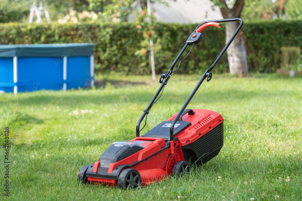 A red lawn mower stands on green grass in a backyard. In the background, a blue pool and hedge are visible. A red lawn mower stands on green grass in a backyard. In the background, a blue pool and hedge are visible.