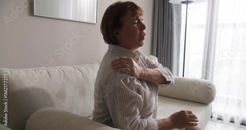 A senior woman with neck pain, gently rubbing the muscles of her neck and shoulder to reduce severe discomfort. An older woman experiencing discomfort in her neck, using her hands to massage the