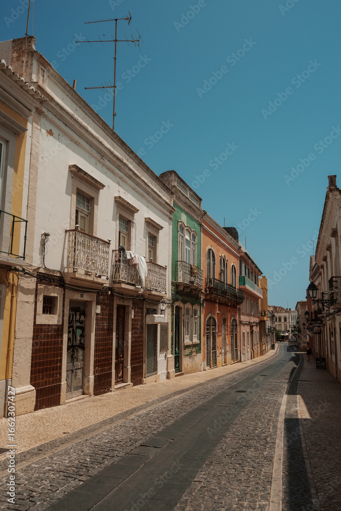 Fototapeta premium Colorful traditional buildings lining a narrow cobblestone street under a clear summer sky in Algarve, Portugal.