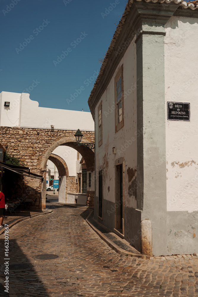 Obraz premium Cobbled street leading to historic stone arches and whitewashed buildings under a clear blue sky in Algarve, Portugal.