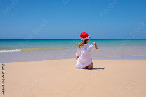 Young girl sitting in red santa hat and white shirt on the ocean shore
