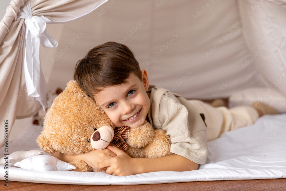 Happy 5 years old little boy lying in a wigwam tent, hugging soft toy teddy bear and looking at camera. Children's room, childhood, games, imagination. High quality photo Happy 5 years old little boy lying in a wigwam tent, hugging soft toy teddy bear and looking at camera. Children's room, childhood, games, imagination. High quality photo