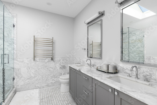 Wall Mural A bathroom with a large grey vanity cabinet, marble tiled walls and flooring, chrome faucets on a white marble countertop, and walk-in shower