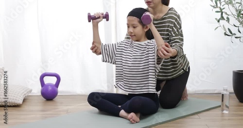 A young girl is training with dumbbells, with her mother helping her. Fit young people lift dumbbells and weights in a home, exercising for a healthy lifestyle concept