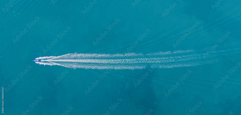 Φωτογραφία Aerial view of a speedboat cutting through clear turquoise sea, leaving a trail behind with anchored cargo ships in the distance