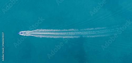Aerial view of a speedboat cutting through clear turquoise sea, leaving a trail behind with anchored cargo ships in the distance. Ideal for marine transport, tourism, and coastal scenes.