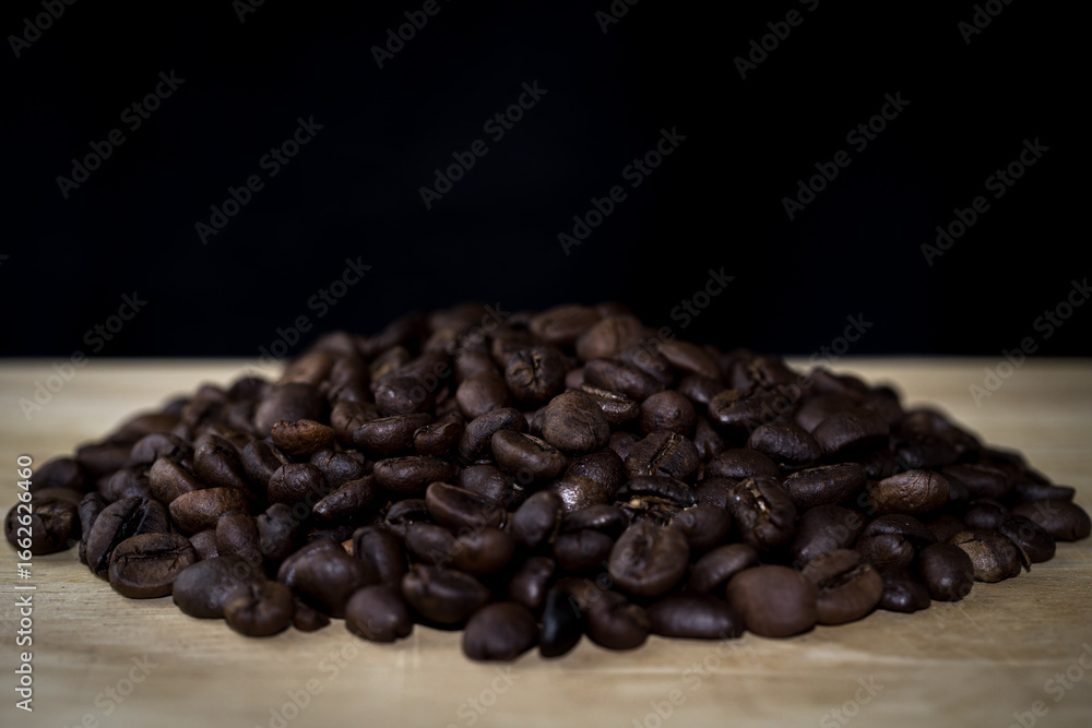 Fototapeta premium A pile of roasted coffee beans placed on a wooden surface with a black background. Perfect for food, beverage, cafe, and product design themes.