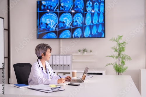 Asian female doctor reviewing brain scans on screen with laptop technology in modern medical office patient diagnosis research healthcare