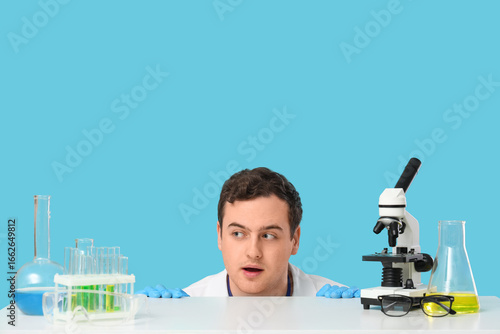 Young male scientist at table with microscope and chemical flasks on blue bac...
