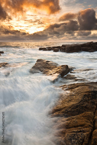 Lorient Morbihan Strong waves breaking over jagged rocks under a dramatic golden sunset.
