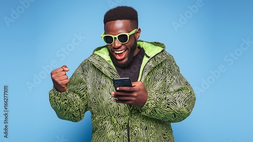 A stylish black guy with a green and black jacket, cool sunglasses, and a smile on his face is clutching his phone excitedly.