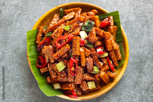 Top view close-up shot of Tempeh or Tempe Orek on a brown plate. A dish featuring stir-fried tempeh strips, coated in a savory sweet sauce, and contrasted with red chilies and green spring onions.