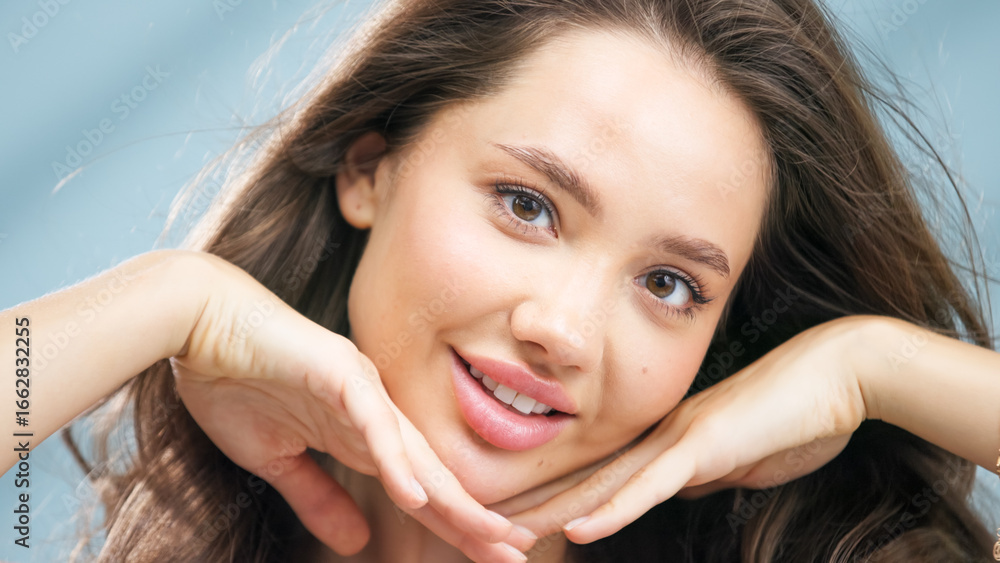 Fototapeta premium Young woman with healthy skin smiling and posing with hands under chin, showcasing radiant complexion and natural beauty, emphasizing skincare and self-care in a bright, inviting atmosphere